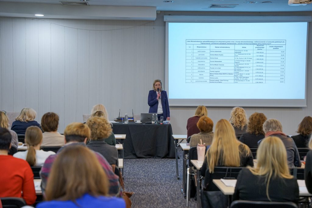 Pełna sala konferencyjna, uczestnicy siedzą w rzędach, tyłem. przodem kobieta mówi do mikrofonu, w tle slajd