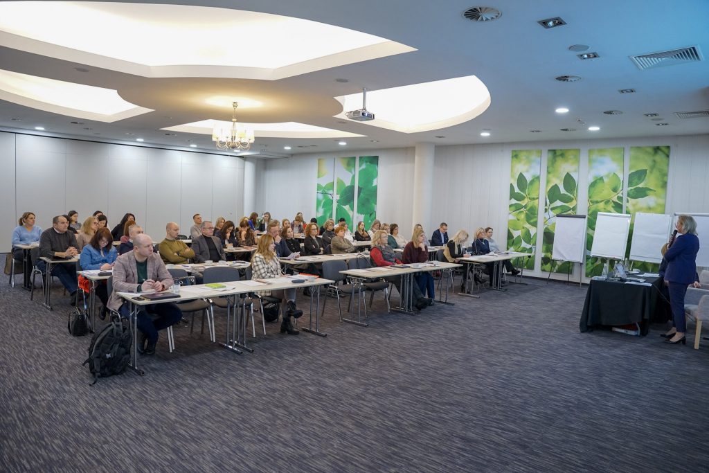 Pełna sala konferencyjna, uczestnicy siedzą w rzędach, z prawej strony prowadząca konferencję kobieta