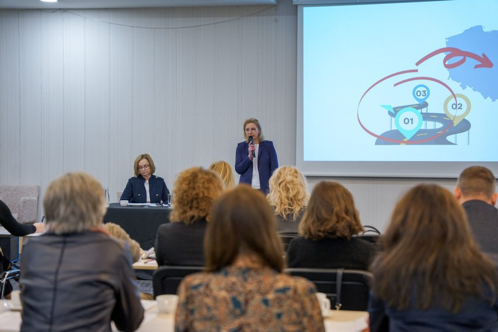 kobieta prowadzi konferencję, w tle slajd. obok siedzi druga kobieta. grupa ludzi pokazana od tyłu
