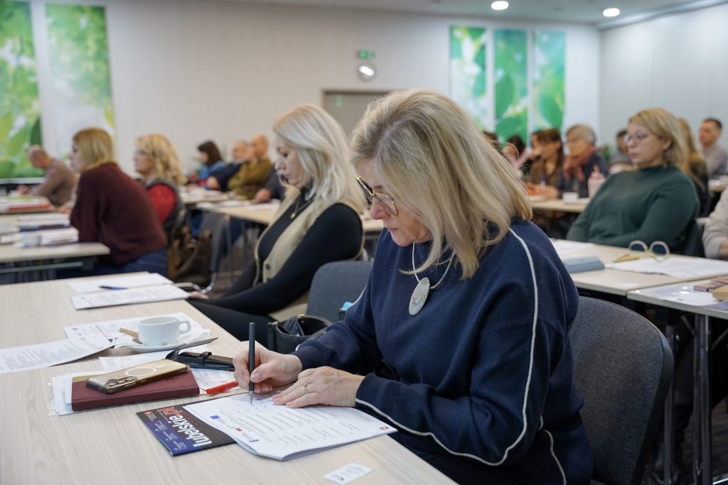 Pełna sala konferencyjna, uczestnicy siedzą w rzędach, kobieta na pierwszym planie notuje