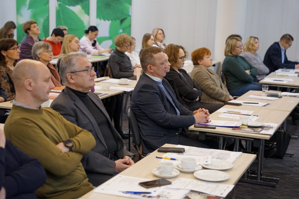 Ludzie siedzą i słuchają konferencji. Na stołach notatki.